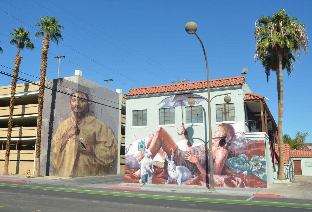 Two large murals on two different buildings that are across the alley from each other. On the left is a large man by Borondo and on the right is a woman reclining on a beach by Finton Magee. She is holding a purple umbrella. There are also cabbages and white rabbits in the scene. 