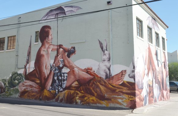 looking at a corner of a two storey white building with a mural painted on the two sides. On the side closest to the camera ia man sitting and holding an umbrella. He is wearing shorts and is topless. A rifle is behind him, white rabbits are also in the picture. 