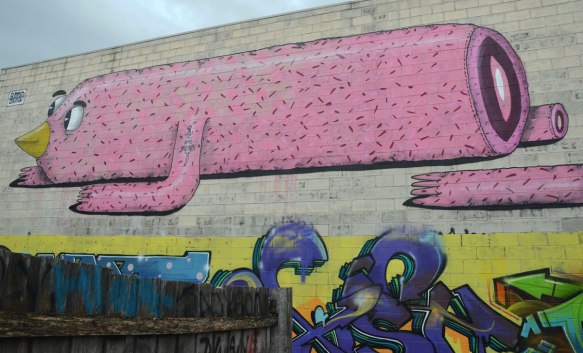 mural on a wall in alley, top part is a long pink animal and the bottom part is a text graffiti tag