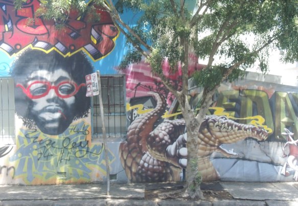 A mural on a wall, includes a black man with beard and mustache and red rimmed glasses, also includes a realistic looking crocodile 