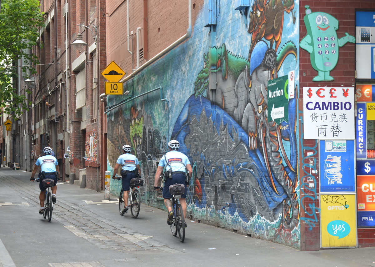 Three policemen on bikes ride down a lane past a street art mural 