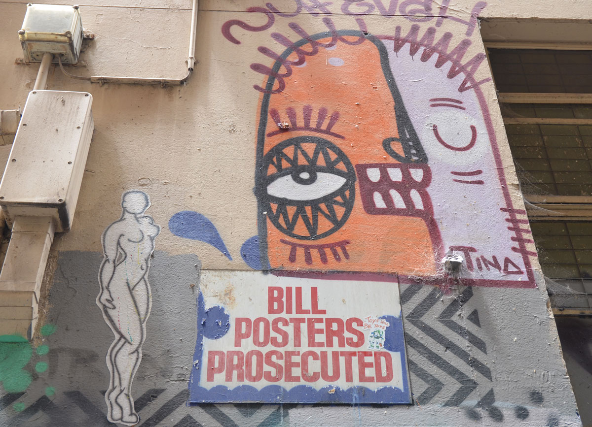 a wall in a laneway in Melbourne, on it are three things, a sign that says "Bill Posters Prosecuted", a paper wheatpaste of a silhouette of a naked woman, and a face in two colours - orange on one side and white on the other, with the name Tina written on it. The orange side has eye open and the white side has eye closed. 