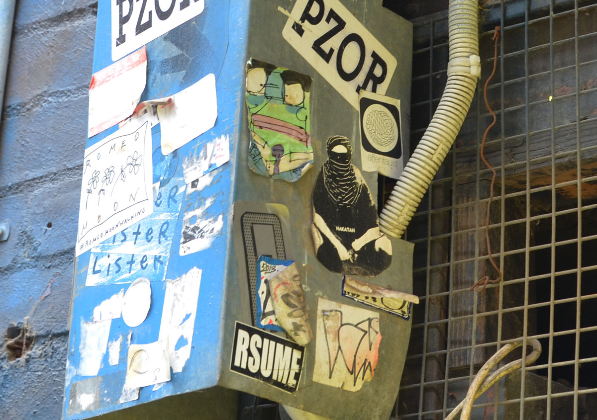 Many slaps and stickers on a metal box in an alley. Stickers with Pzor written on them and sticker saying Rsume. A picture of a person in black robes and a veil. 