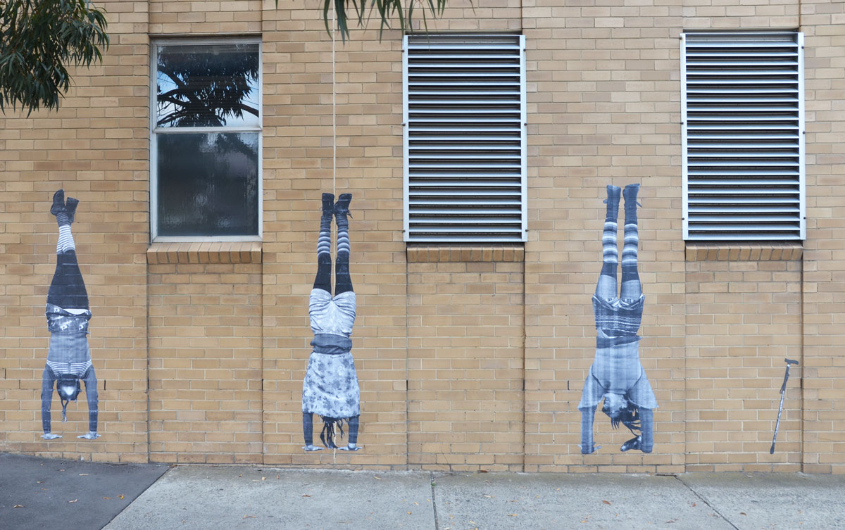 street art paste ups of three people standing on their hands, with a cane to the right of them. 