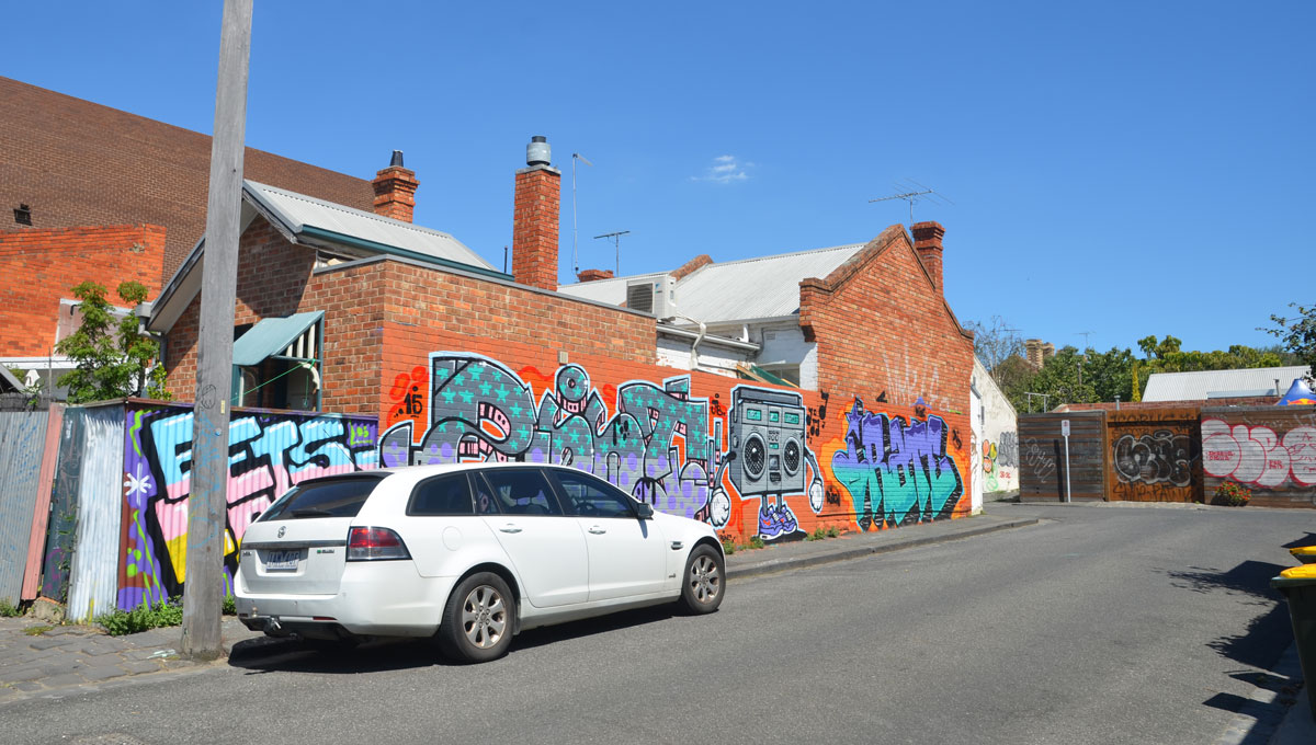 a white car is parked on a street in front of a long wall with street art on it, two tags, or text graffiti and a ghetto blaster painting. 