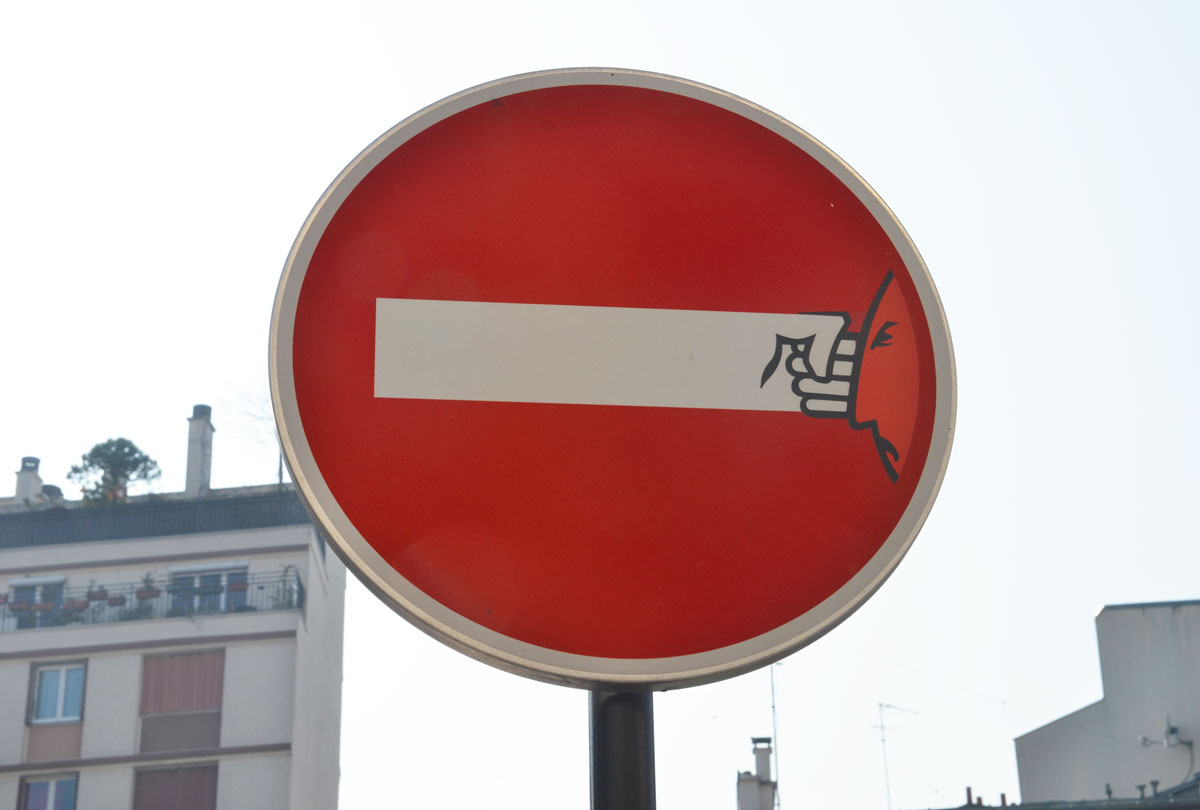 graffiti altered no entry sign, red circle with a white horizontal bar - added face on right side, lines on bar to make it look like an arm and a fist, punching the face