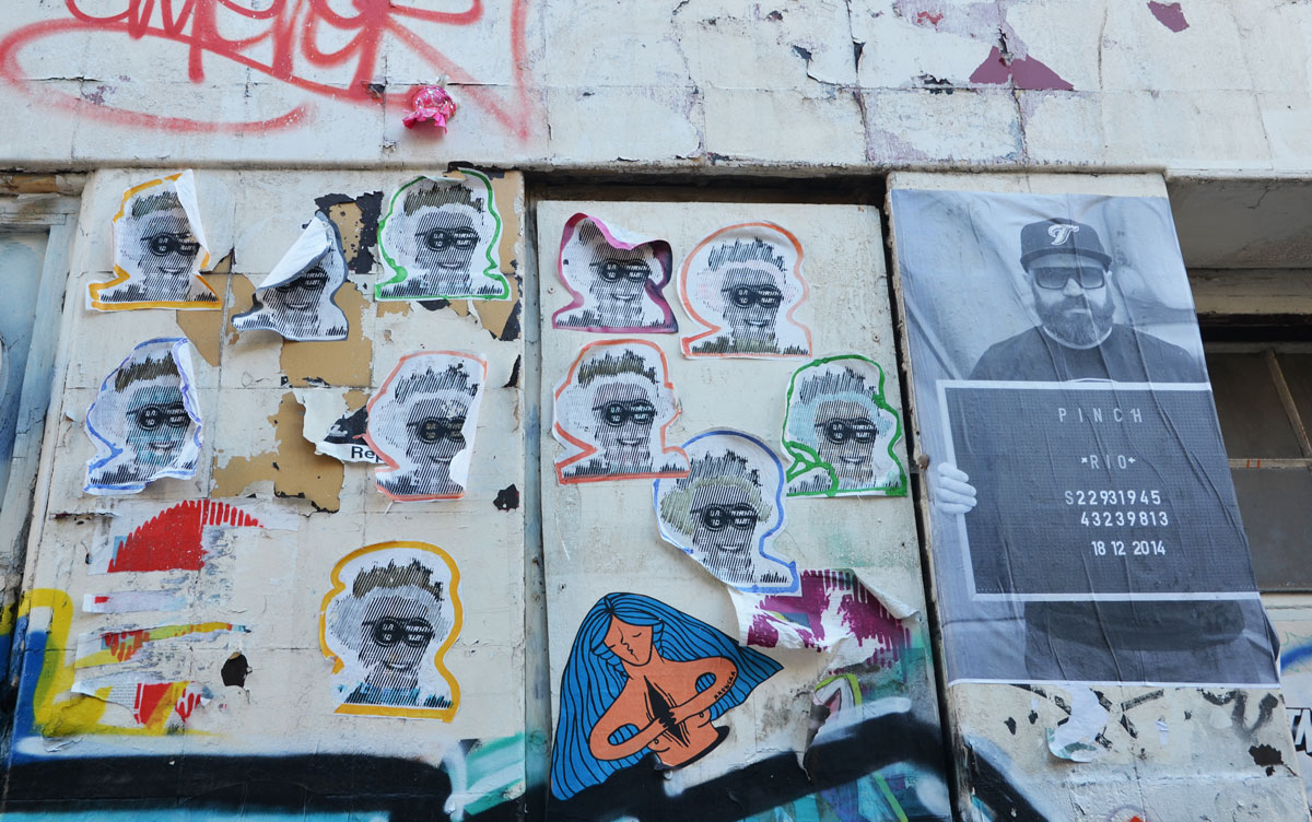 many identical paste ups of Queen Elizabeth's head, outlined in different colours, pasted on a wall, 
