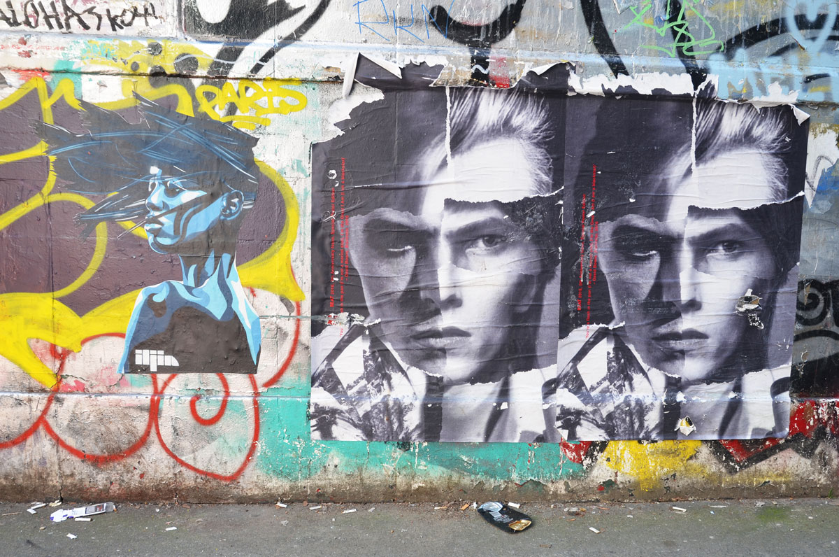 three graffiti faces on a wall, one woman in profile painted in blues and two identical black and white paste ups of a young man 