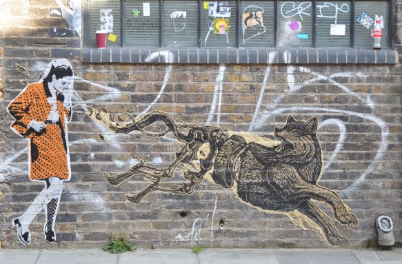 Two paste ups on a wall, a running wolf with hind legs outstretched to the back, and a woman in an orange and black outfit 