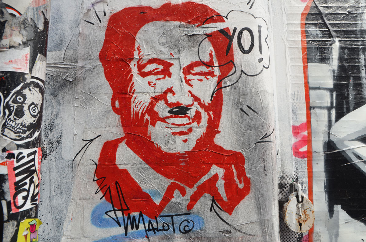 a man's head, stencil in red, someone else has given him a black Hitler moustache and a word bubble that says yo! Signed malot. 
