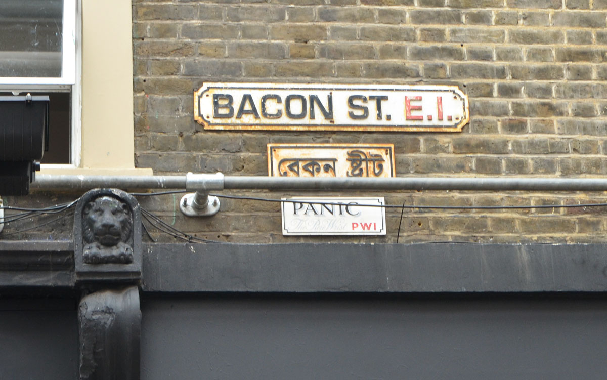 London street sign, Bacon St., E.1 with its Arabic counterpart below it. Also, a PANIC sign that is supposed to look like a street sign under that. 