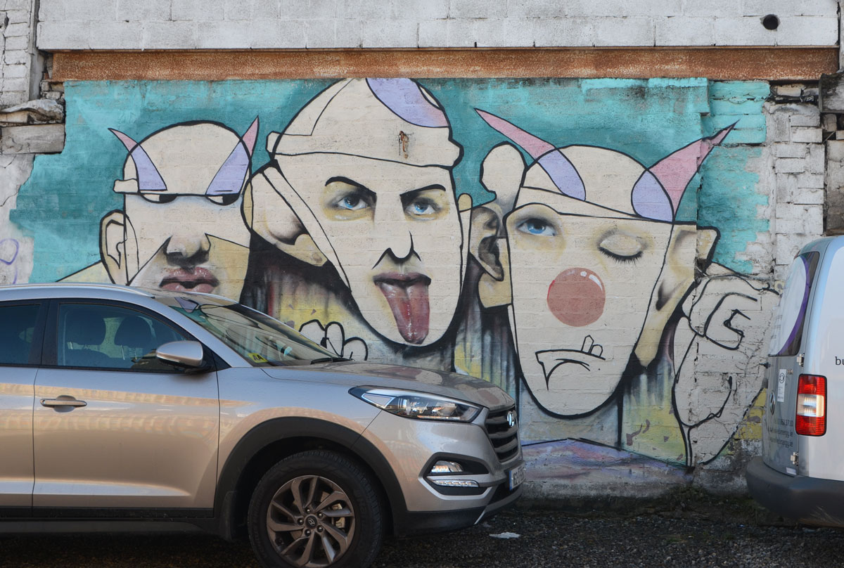street art of three male faces, jester like hats, in subdued tones of blue and pink and beige . Line drawings but with realistic lips and tongue