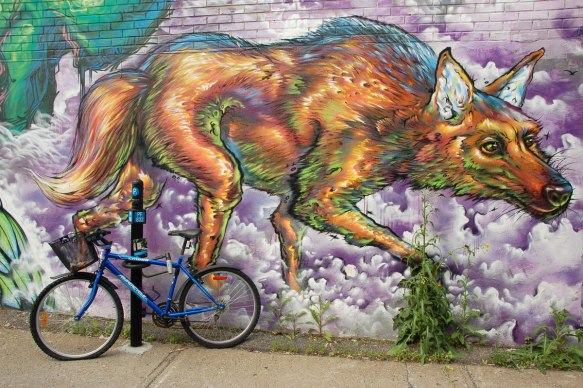 street art, graffiti, animals painted on a wall - an ugly awkward looking dog paces in a mural. The dog looks like it's walking on the sidewalk. A bike is parked in front of it