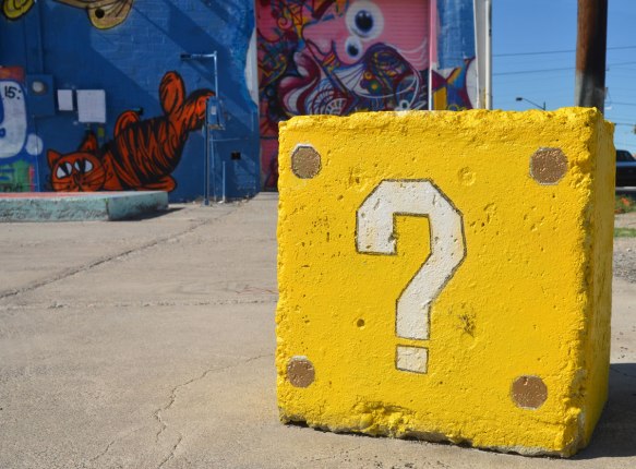 white question mark on a concrete block painted yellow, with a mural covered on a wall in the background