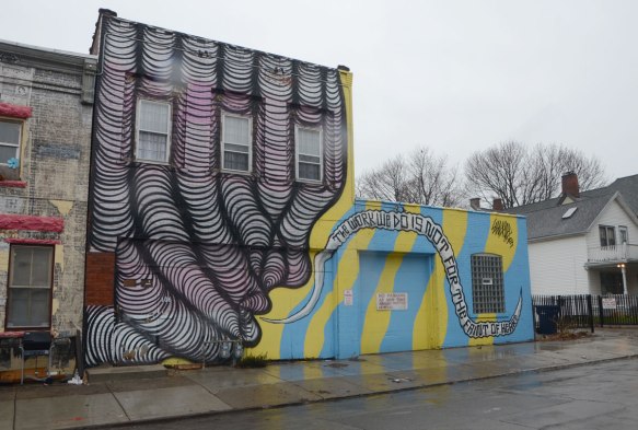 a mural on the side of a building. On the left is a purple and black drawing of what looks like fingers. on the right are yellow and turquoise stripes with the words, the work we do is not for the faint of heart