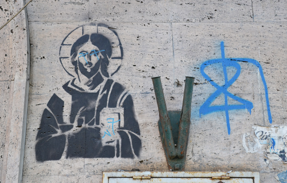 black stencil of Jesus, with halo around his head, a book in one hand, the other hand is pointing the book 