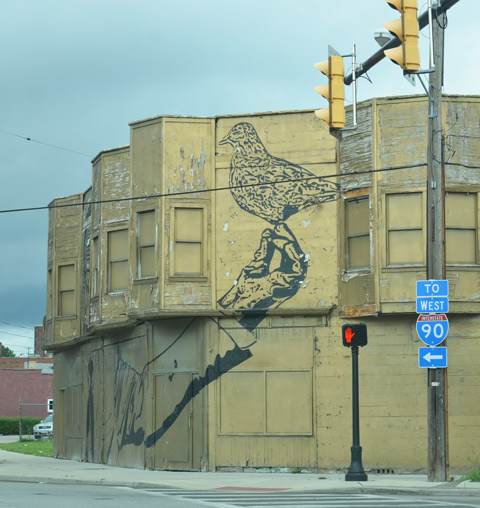 continuation of mural of man in gas mask, building is on a corner and the mural exte4nds around the corner, there is a large bird in his hand, 