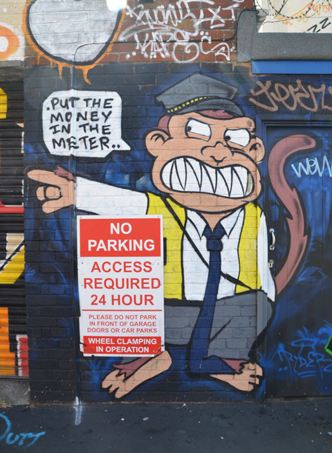 parking meter man as a monkey, sneering, and pointing, beside a sign that says no parking, words on street art say put the oney in the meter