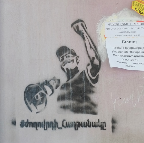 black stencil of a man holding a megaphone in one hand and with the other hand raised in a fist, words in Armenian written below it. 