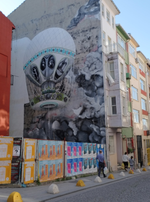 large mural in Kadikoy on the side of a building, hot air balloon, on the side of a building, vacant lot beside behind hoardings covered with ad posters Painted by Brazilian artist Claude Ethos