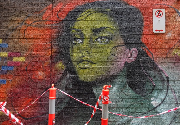 orange and white traffic markers in front of a mural in a lane, on a brick wall, of a woman's face and head, green skin on face, blue skin on neck, long black hair, blue eyes, 3/4 profile position 