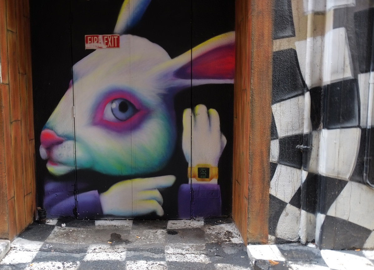 mural of a white rabbit from Alice in Wonderland, with pinkish eyes and ears, and purple jacket, and wearing a wrist watch, on a double door, surrounded by black and white checked pattern. 