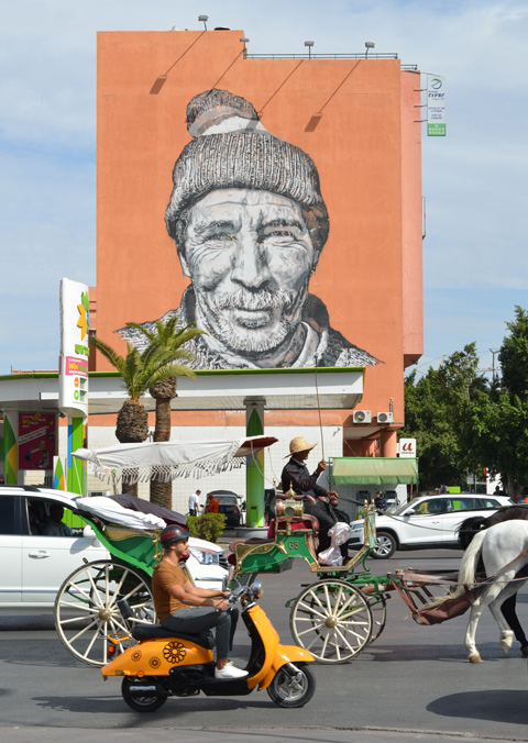 traffic passing by including a horse drawn carriage, a gas station on the corner across the street and a large mural of a berber man on a wall. Mural by Heinrich 