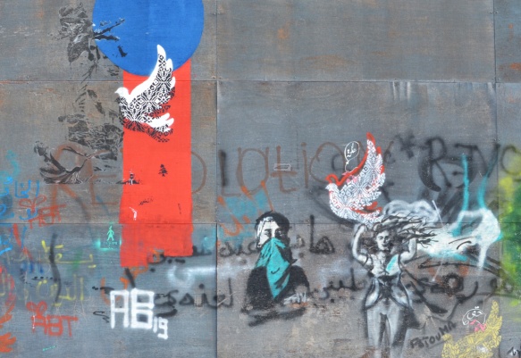 blue circle, red vertical stripe, black stencil of doves and other things in black along with a woman with a teal coloured bandana over the lower part of her face, a woman with long hair trying to hold onto a dove, all stencils 