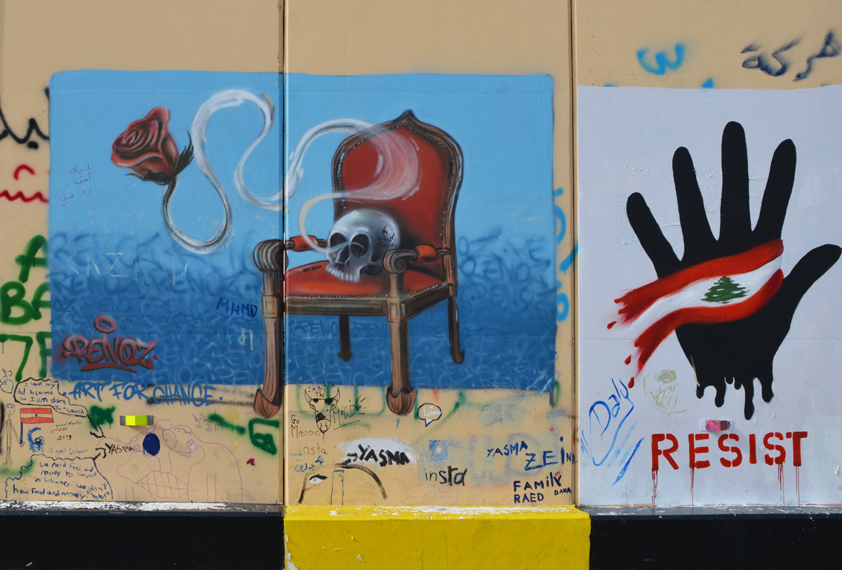 a skull sits on a red upholstered chair with wood arms and legs, on a blue background, also hand with lebanese flag, and the word resist 
