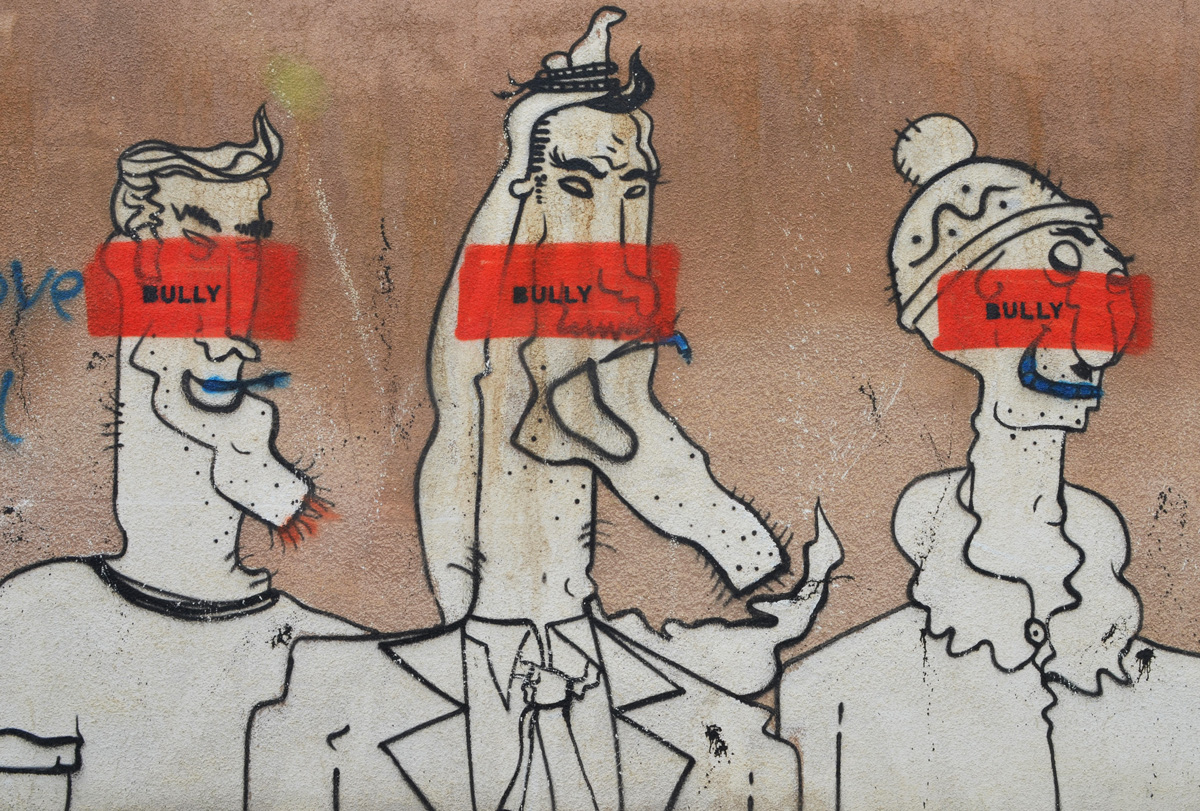 on a brown wall, three comical white characters, male, with exaggerated features drawn in black, each has an orange stripe through their face with the word Bully written on it 