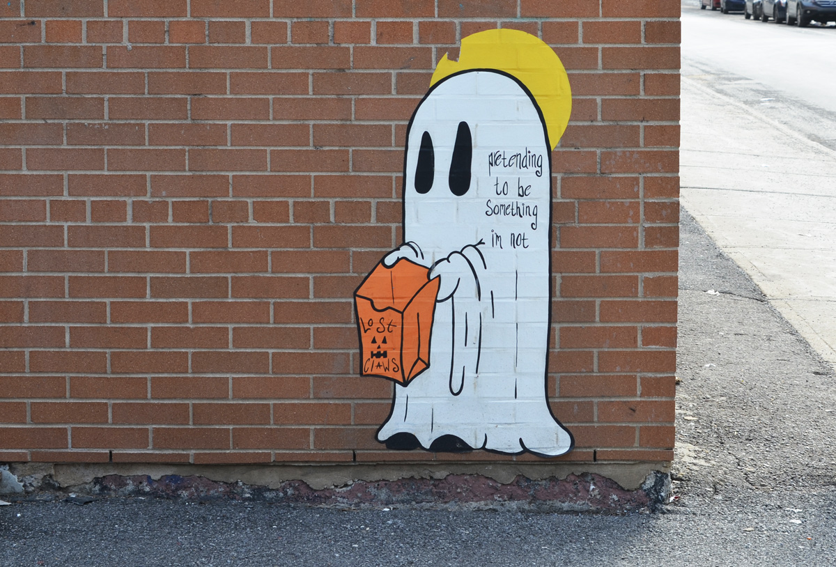 paper pasteup of a ghost costume, white sheet, with black eyes, also little hands holding an orange bag. words on the ghost saying pretending to be something i'm not 