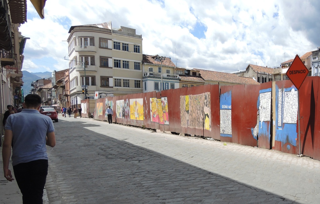 a man walking down a street in Cuenca Ecuador with hoardings surrounding a construction site on one side of the street, the hoardings have murals painted on them