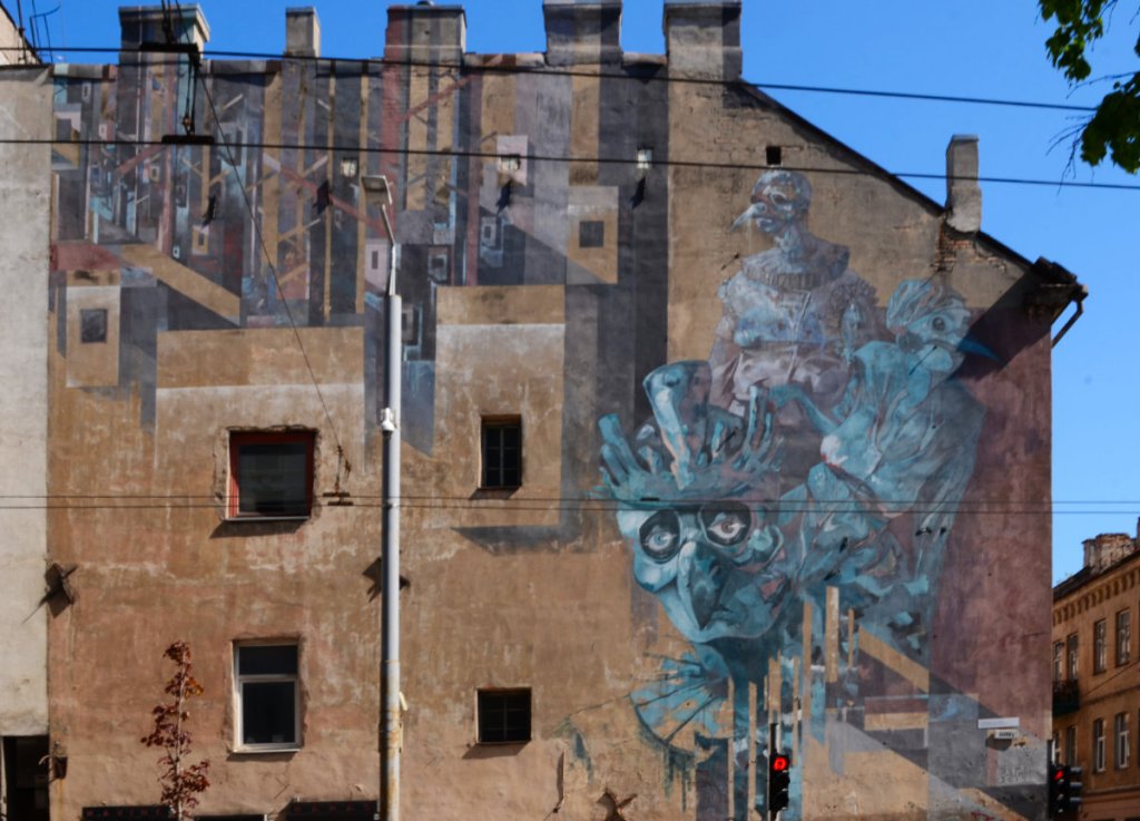large mural on the side of a building, shades of blue and purple mostly, some abstract geometric shapes, 2 figures that are part person and part bird, especially with beaks.  painted by two artists, sepe and chazme