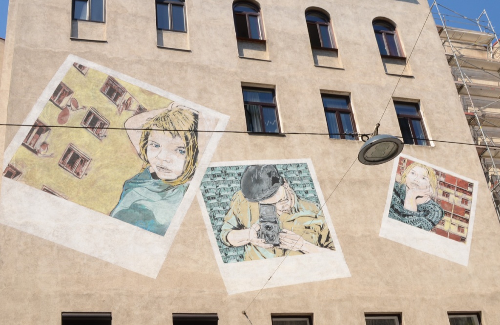 mural on a wall that looks like 3 square polaroid photos. Two are of young women with blond hair standing in front of apartment blocks, and the third is a man who is also taking a polaroid picture 