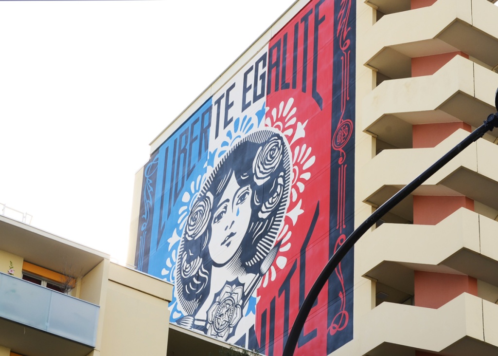 mural by Shepard Fairey in Paris, on top of a building, French motto Liberte, Egalite, Fraternite forming a large circle, with a woman's head in the middle.  Woman has roses in her hair. 