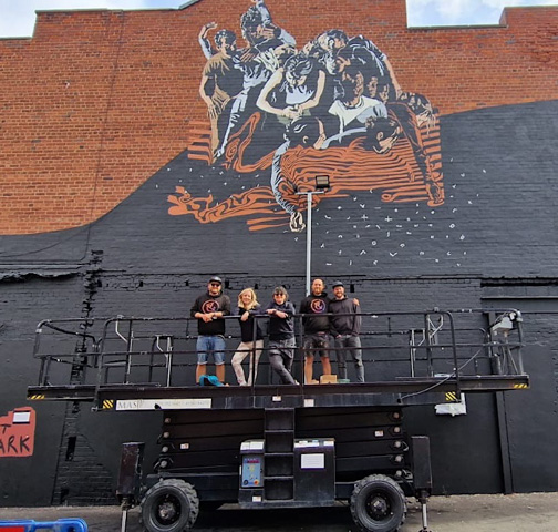 a group of artists posing together on a lift that has been used to paint a mural high on a brick wall