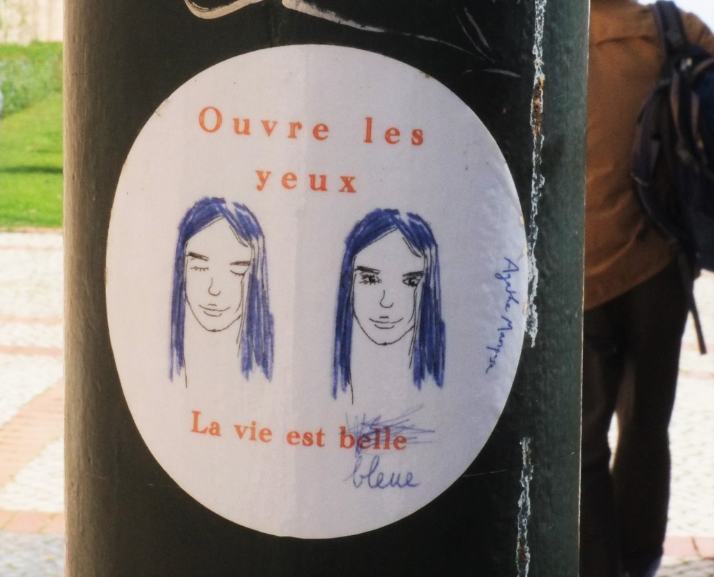 sticker graffiti, two faces, words that say Ouvre les yeux. La vie est belle but someone has crossed out belle and written bleu