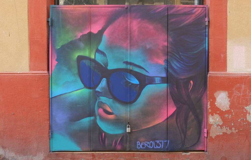 street art mural on metal shutters over windows of Akoda Taberna on Carrer de Blesa, painting of a woman's portrait in blues and purples, wearing dark blue sunglasses, by Berol377