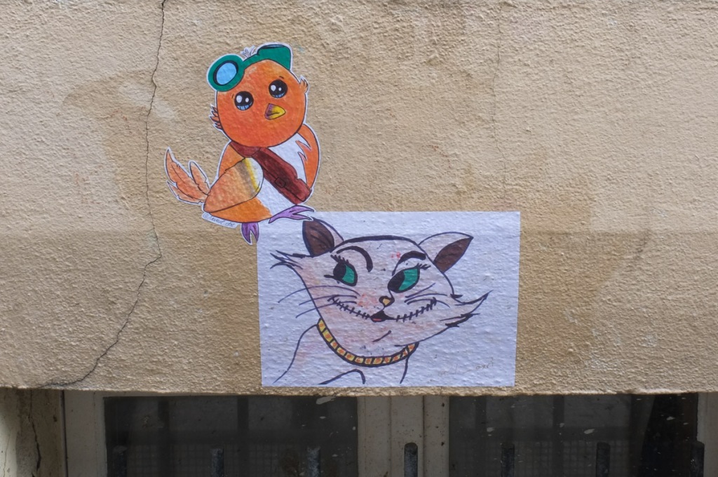 two small wheatpaste graffiti on a wall above a window. One is the head of a white cat, the other is a yellow and orange bird