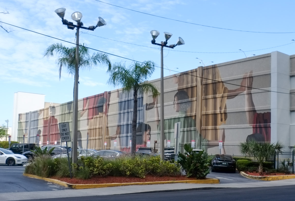 large mural on side of building by parking lot, title is communidad, large people reaching out to one another