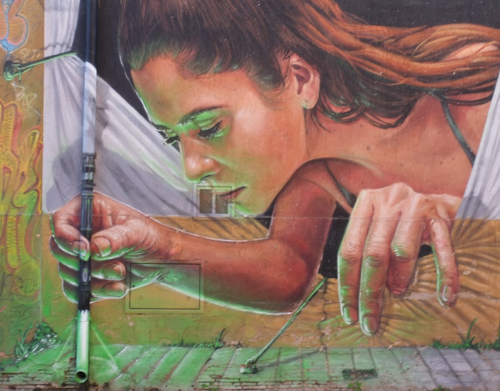 mural on the side of a building of a woman leaning out of a window with a white curtain, holding a pen (that is in reality a drain pipe) and writing on the sidewalk, by French artist Braga Last one
