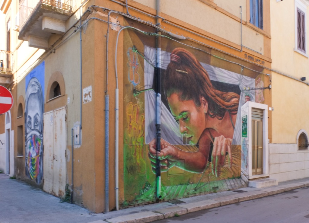 one one side of a corner is a mural painted in 2020 of close up of a woman's face, wearing a brightly coloured covid mask and on the other side is a mural of a woman leaning out of a window, both in Stornara Italy