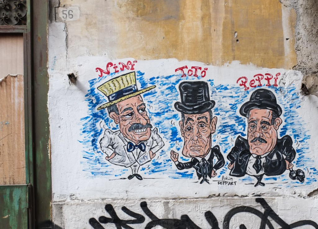 caricature drawings, paper paste up on an alley wall, of three older Italian actors, with stage names, Toto, Nini, and Peppi