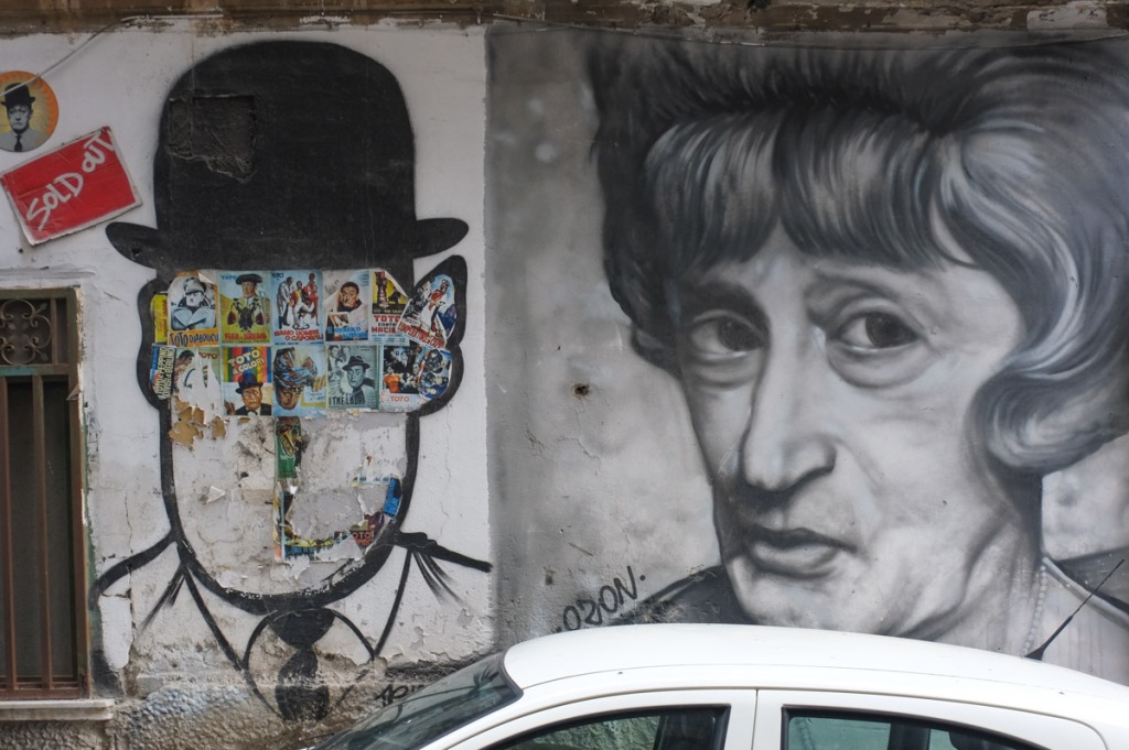 two street art pieces, a black and white painting of a man in drag, a black line drawing of man in a black bowler hat but the face is lost, and stickers have been put there instead