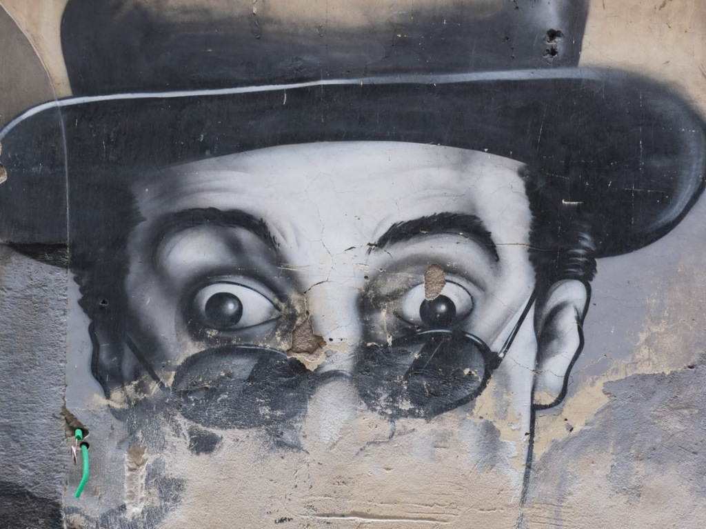 black and white mural, portrait of actor Toto in sunglasses at end of his nose, wearing black bowler, lower part of mural has been worn away