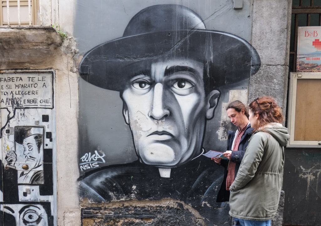 a couple stands in front of a street art painting of an actor dressed as a Roman Catholic priest in a black and hat and white clerical collar